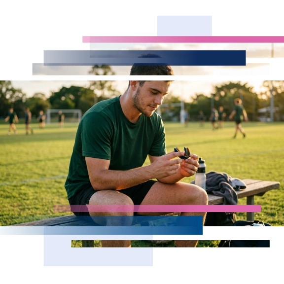 Player sitting on a bench, holding a mouthguard, with a grassy field and players in the background during sunset.