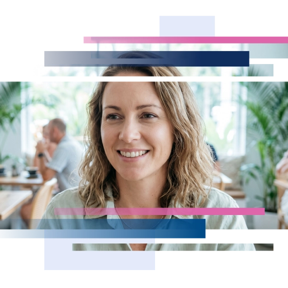 Smiling woman with wavy hair sitting in a cafe with plants and a blurred background.