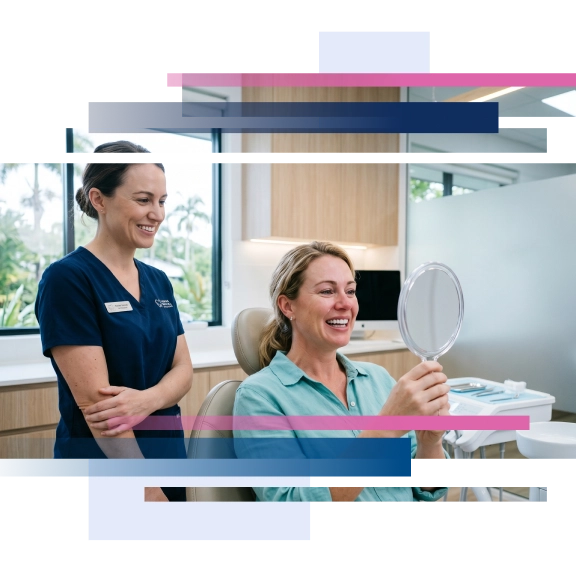 Woman smiling while holding a mirror in a dental office, with a staff member standing beside her.